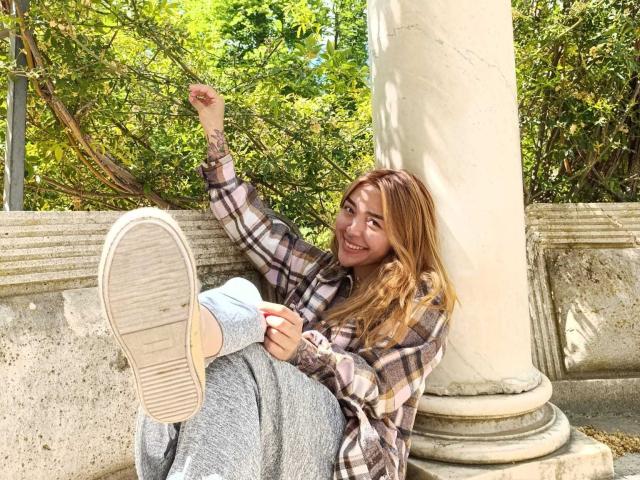 SeleneAna with long blonde hair, smiling, wearing a plaid shirt and grey sweatpants, legs up, showing a sole of a sneaker, arm tattooed, leaning against a white column.