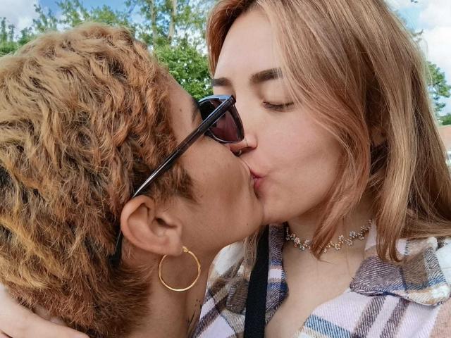 SeleneAna with curly brown hair kissing another SeleneAna with straight blonde hair, wearing sunglasses and a choker. Both have hoop earrings and are kissing passionately.
