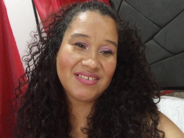 GotitaCeleste hat lockiges, schwarzes Haar, braune Haut, rosigen Lidschatten, rosigen Lippenstift und lächelt mit einem leicht rundlichen Gesicht, wobei ihre Zähne sichtbar sind. Sie trägt keine Kleidung..
