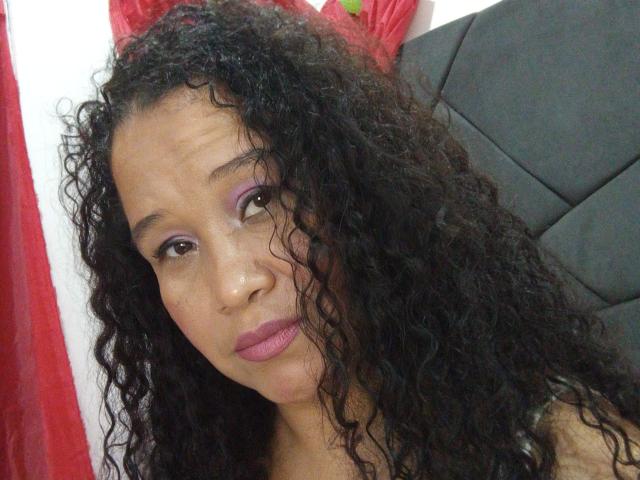 GotitaCeleste tem cabelo preto encaracolado, olhos castanhos, sombra roxa, batom rosa e uma expressão neutra. Ela está sentada em uma cadeira preta, com cortinas vermelhas ao fundo..