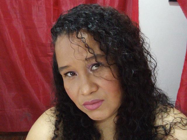 GotitaCeleste tiene el cabello oscuro y rizado, ojos marrones y lápiz labial rosa. Se puede ver su rostro ligeramente fruncido y sus hombros desnudos. En el fondo, hay una cortina roja..