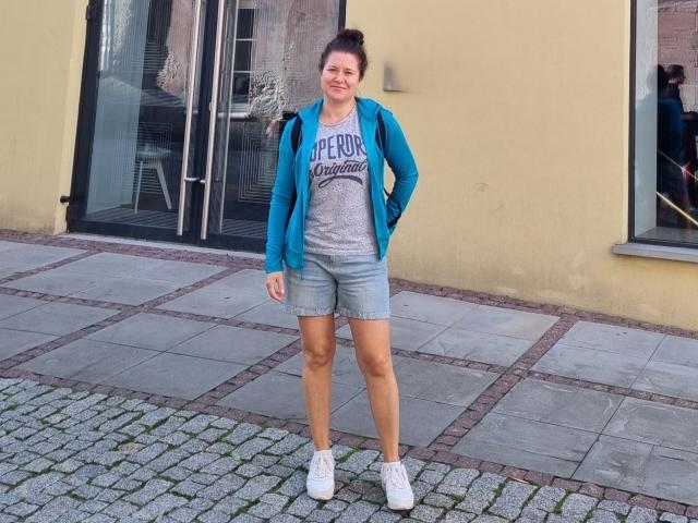 FlirtYolivia, mit einem Lächeln, trägt eine blaue Jacke, ein graues Hemd, Jeansshorts und weiße Turnschuhe. Sie steht auf einem Kopfsteinpflasterweg, die Hände in den Taschen, mit einem Rucksack auf dem Rücken, und hat braunes Haar zu einem Dutt gebunden..