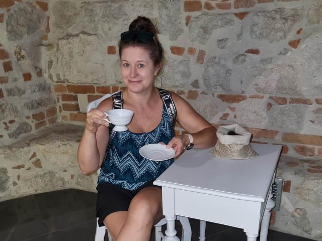 FlirtYolivia with tits in a blue patterned top, black shorts, holding a white cup and saucer, sitting at a white table with a tissue box.