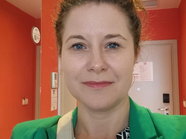 FlirtYolivia con ojos azules, lápiz labial rosa, camisa verde, correa blanca y pared rojo-naranja, sonriendo ligeramente, cabello castaño en un moño despeinado, piel clara y un rostro lindo y relajado..