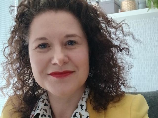 FlirtYolivia mit lockigem, braunem Haar, rotem Lippenstift und einer gelben Jacke, lächelt leicht und trägt einen weißen und schwarzen Schal mit Muster. Sie hat blaue Augen und einen hellen Hautton..
