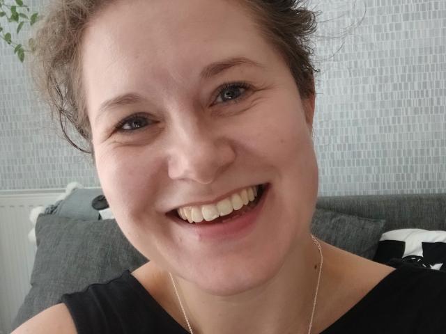 FlirtYolivia, com um grande sorriso feliz, mostrando os dentes, e vestindo uma blusa preta. Ela tem cabelo castanho encaracolado, pele clara e um colar de prata. Seus olhos são azuis..