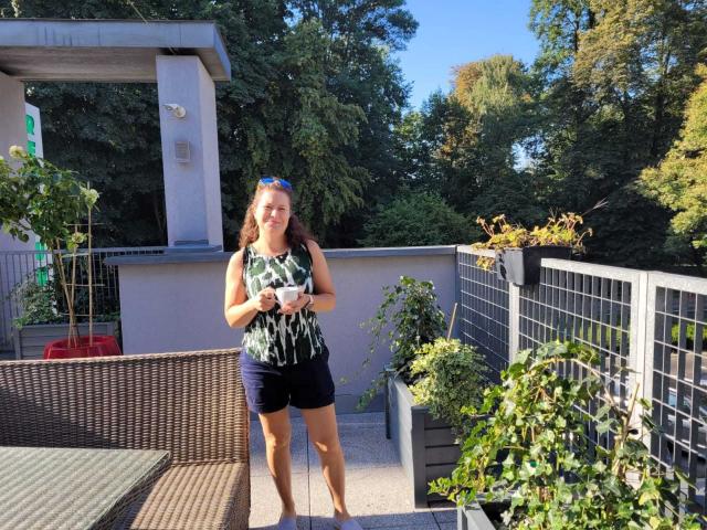 FlirtYolivia mit Hintern in dunkelblauen Shorts, Oberteil mit grün-schwarzem Muster, hält eine weiße Tasse, steht auf einer Terrasse, mit einem Korbstuhl, trägt blaue Sonnenbrille, im Hintergrund Bäume und Topfpflanzen..