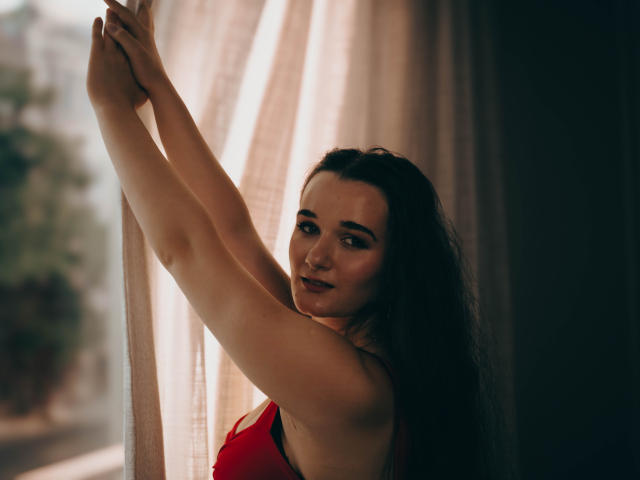 KateUnique with dark hair, arms raised, looking over a white surface, smiling subtly, holding a curtain, her tits slightly visible, soft lighting, warm brown eyes.