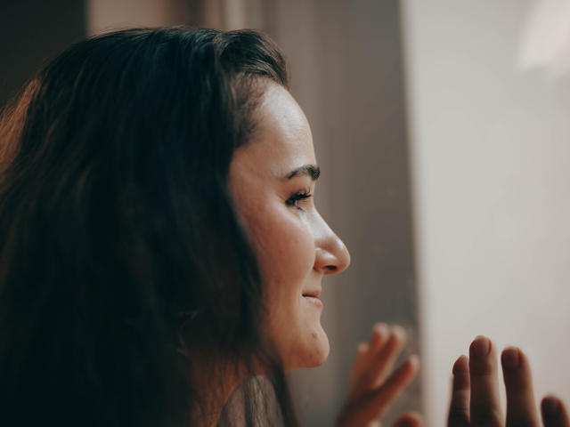 KateUnique, com cabelos castanho-escuros ondulados, sorrindo suavemente, olhos fechados, mãos levantadas, luz suave no rosto, maquiagem discreta, relaxada e feliz, leve rubor nas bochechas..
