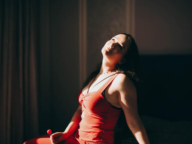 KateUnique, avec une poitrine de taille moyenne, portant une robe rouge, est assise, les yeux fermés, la tête inclinée, avec des cheveux foncés, un léger ombrage, une lumière douce, une expression détendue et sensuelle, sur un fond sombre..