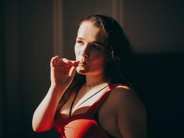 KateUnique, avec ses cheveux bruns mouillés, sa robe rouge et son collier, mordant sa lèvre, la lumière du soleil mettant en valeur ses seins et son visage, fond noir, regard séduisant, tenant le collier dans sa main..