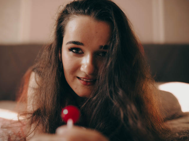 KateUnique, avec de longs cheveux bruns ondulés, tenant un vibromasseur rouge, souriant de manière séduisante à la caméra, ses seins partiellement visibles, allongée sur un lit, lumière douce, ambiance intime..