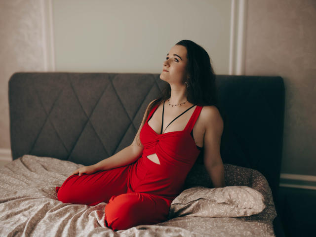 KateUnique, met een rode jurk en uitsparingen, zit op bed, kijkt omhoog, haar borsten zijn licht zichtbaar, donker haar, ketting, armen rusten, benen gekruist, zacht licht, ontspannen..