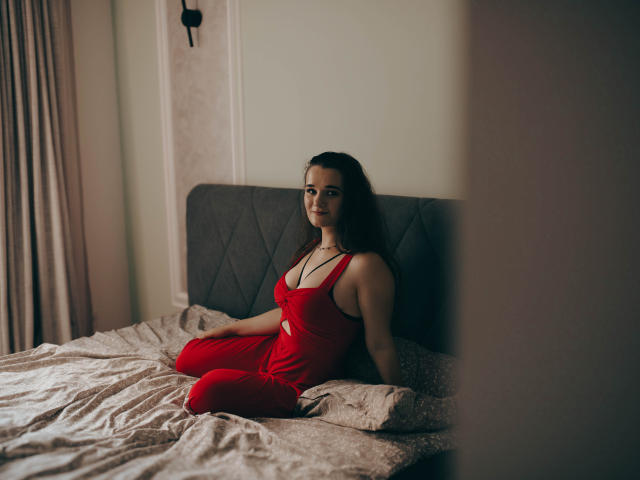 KateUnique, sitting on a bed, wearing a red dress that shows her tits and a cutout, with long dark hair, looking slightly annoyed, in a dimly lit room.