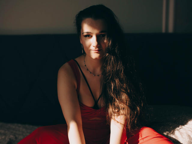 KateUnique, con cabello largo y castaño, vestido rojo y sujetador negro, sentada en la sombra, con luz en el rostro, mirando a la cámara, con un collar pequeño, fondo oscuro, con una sonrisa sutil..