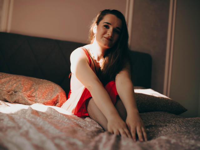 KateUnique sitzt auf einem Bett, trägt ein rotes Kleid und Fischernetzstrümpfe, die Arme sind über den Knien verschränkt, sie lächelt in die Kamera, das Sonnenlicht betont ihr braunes Haar und ihre helle Haut..