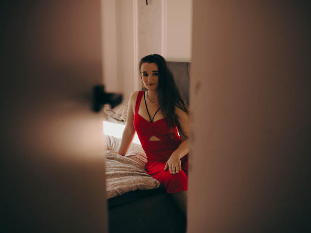 KateUnique in a red dress, sitting on a bed, looking at the camera, with long brown hair, small tits, and a subtle smile, through a partially open door.