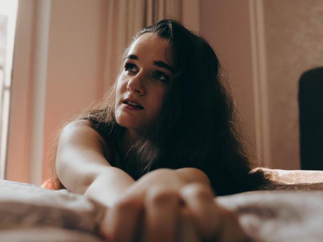 KateUnique with long dark hair, lying on bed, reaching forward, soft light, pinkish skin, slightly parted lips, looking upwards, subtle smile, outstretched arms, cozy indoor setting.