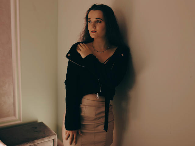 KateUnique, con cabello largo y oscuro, vestida con una chaqueta negra y una falda beige, está de pie contra una pared, con una mano en el pecho, mirando hacia un lado, mostrando sus pequeños pechos y glúteos..