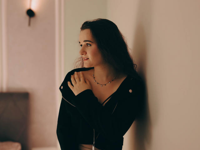 KateUnique with long dark hair, wearing a black top, touching her collarbone, smiling softly, necklace, wall light in background, warm light, subtle shadow, casual, attractive.