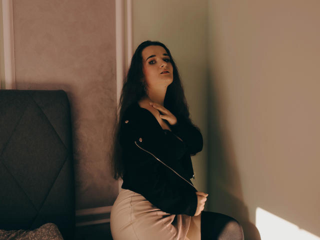KateUnique, con lunghi capelli scuri, giacca nera e gonna beige, seduta, con una mano sul petto, bocca leggermente aperta, sguardo seducente, luce soffusa, testiera grigia, ombre calde..