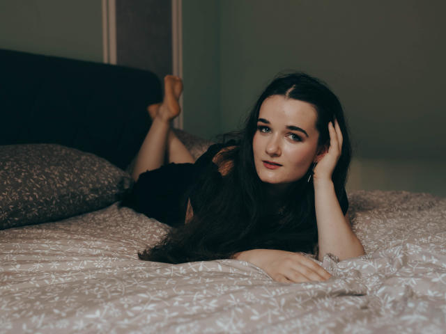 KateUnique liegt auf einem Bett, die Beine hochgelegt, trägt ein schwarzes Oberteil, hat langes, dunkles Haar, blaue Augen und helle Haut, ihre Hände liegen auf dem Kopf, und sie schaut in die Kamera..