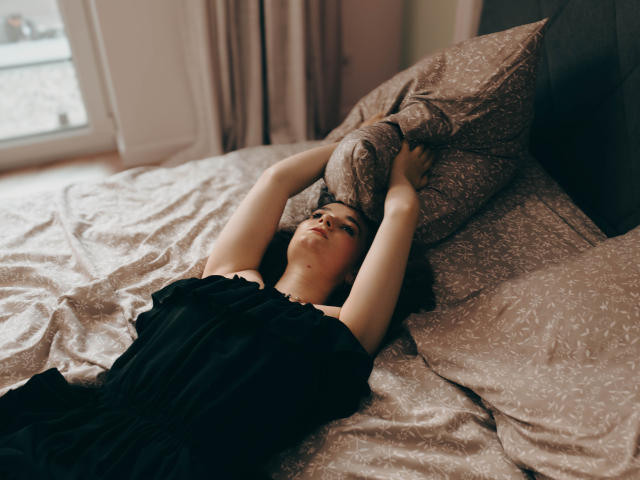 KateUnique ligt op een bed met haar armen boven haar hoofd, ze draagt een zwarte jurk en haar handen houden een gestreept kussen vast. Ze kijkt omhoog met een licht geïrriteerde uitdrukking..