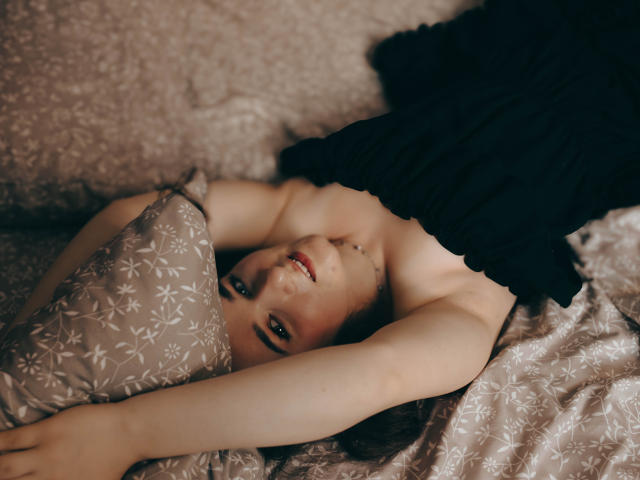 KateUnique est allongée sur un lit, les bras au-dessus de la tête, portant un haut noir, avec ses seins légèrement visibles, et un oreiller fleuri sous son visage..
