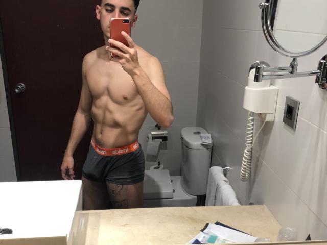 MiguelHt, shirtless, muscular, wearing grey Calvin Klein underwear, holding red phone, taking mirror selfie, visible tattoo on right thigh, white bathroom, toilet, wall-mounted toilet paper holder.
