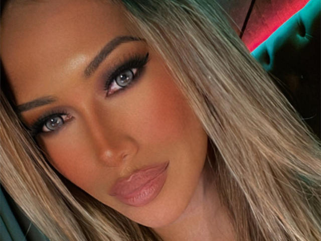 JasMully's face, featuring blue eyes, thick eyebrows, long blonde hair, and pink lips, with a slight smile and subtle makeup. Her tits are not visible.
