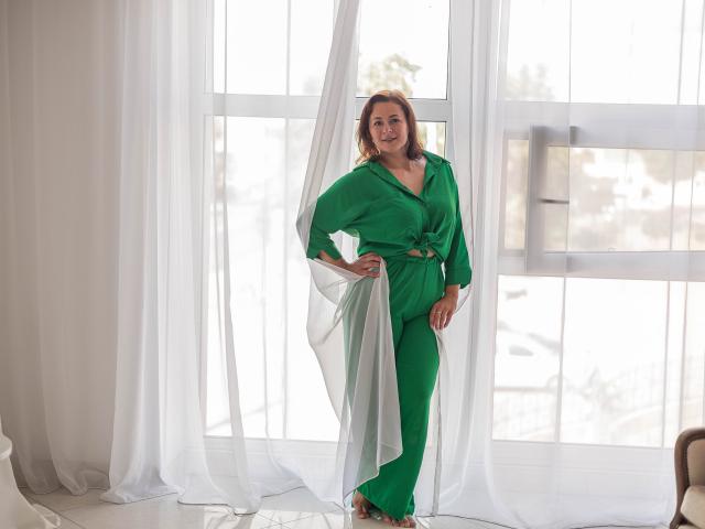MarieSho está de pie con un atuendo verde que incluye mangas amplias y pantalones, sonriendo con las manos en las caderas, cerca de cortinas blancas y una ventana iluminada por el sol detrás de ella..