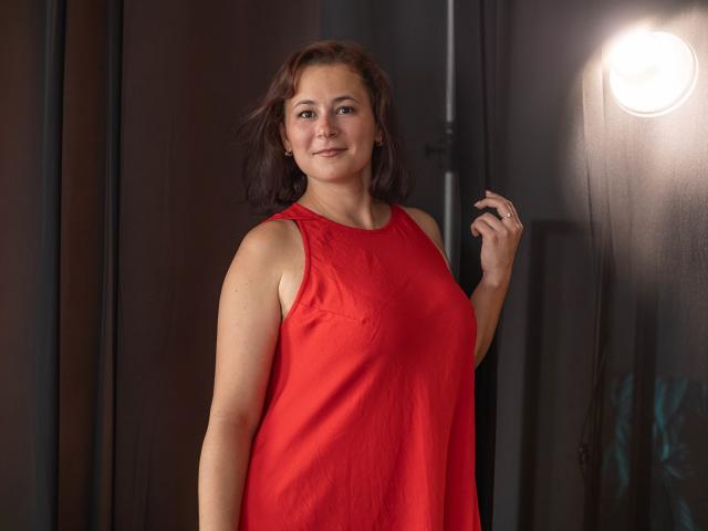 MarieSho with medium tits, wearing a red sleeveless top, standing, smiling, right hand up, brown wavy hair, light skin, subtle makeup, soft light from right, dark background.