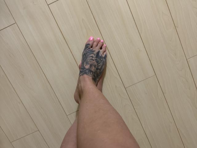 AndraD's foot with a detailed black tattoo on the sole, pink-painted toenails, and her leg, standing on light wooden floor.