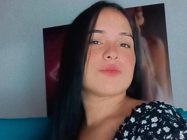 MartinaDupont with black hair, pouty lips, and light skin, wearing a floral top, has a subtle, relaxed expression, and a blurred, dark-colored photo behind her.