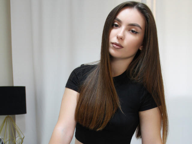 EmmaBettis with long brown hair, wearing a black shirt, has a calm expression, slightly tilted head, and subtle makeup, standing in a room with a black lamp.