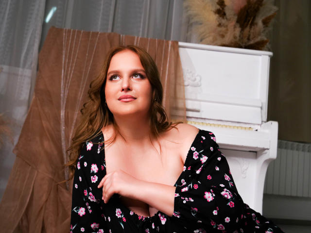 ShiningSmile with wavy brown hair, blue eyes, and a cute smile, wearing a black floral dress, arms crossed, standing in front of a white piano. Boobs visible, soft light.