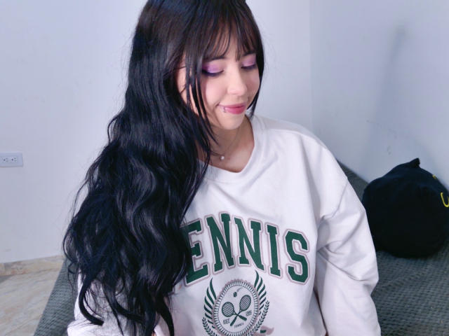 Arieyaara mit langem, schwarzem, gewellten Haar, rosa Lidschatten und einem weißen "TENNIS"-Pullover, lächelt sanft. Ihre kleinen Brüste sind leicht unter dem Shirt sichtbar..