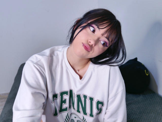 Arieyaara with black hair, purple eyeshadow, white "TENNIS" sweatshirt, and pink lipstick, leaning forward, looking up with a slightly mischievous smile, and a small nose piercing.