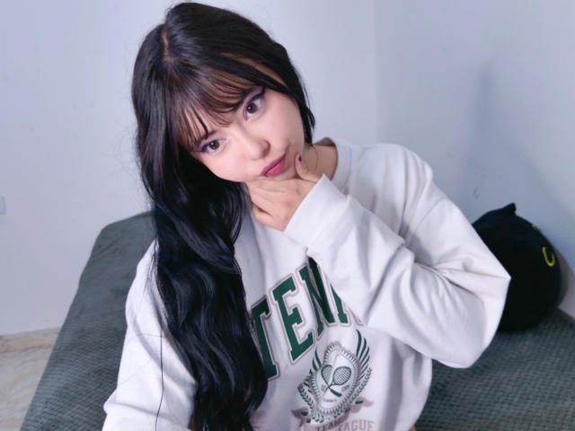 Arieyaara with long black hair, bangs, wearing a white Teem sweater, holding her neck, looking down with a slight smile, sitting on a green bed.