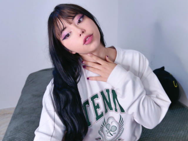 Arieyaara met lang zwart haar, roze oogmake-up en roze lippenstift, draagt een witte GENA hoodie, raakt haar nek aan met handen met roze nagels en kijkt naar de camera..