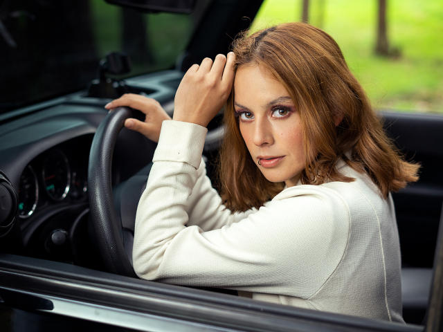 VictoriaOrtega with brown hair, white blouse, and serious expression, hands on steering wheel, inside car, looking back with slight smirk. Her tits are not visible.