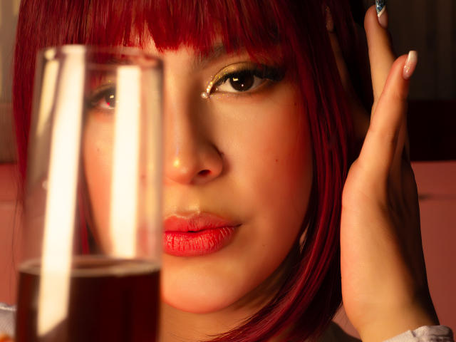 SamanthaEva69 with red hair, glossy pink lips, and gold eye makeup, holding a glass in the foreground, her hand near her ear. Her nails are manicured.