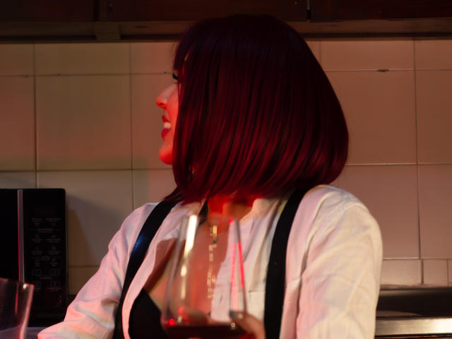SamanthaEva69 with red bob, white shirt, black suspenders, holding a glass, red light on her face, kitchen background, smiling sideways.