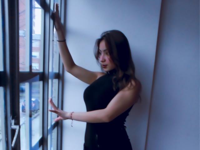 Ashley Miller, avec des seins, portant un haut noir sans manches, debout près d'une fenêtre, les bras levés, regardant à l'extérieur, cheveux bruns ondulés, yeux bleus, maquillage discret, lumière douce, mur blanc..