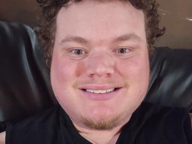 Dillondiape69 hat ein rundes Gesicht mit lockigem, braunem Haar, helle Haut, einen kleinen Bart und ein Lächeln, bei dem weiße Zähne zu sehen sind. Er trägt ein schwarzes Hemd und sitzt auf einem schwarzen Ledersessel..