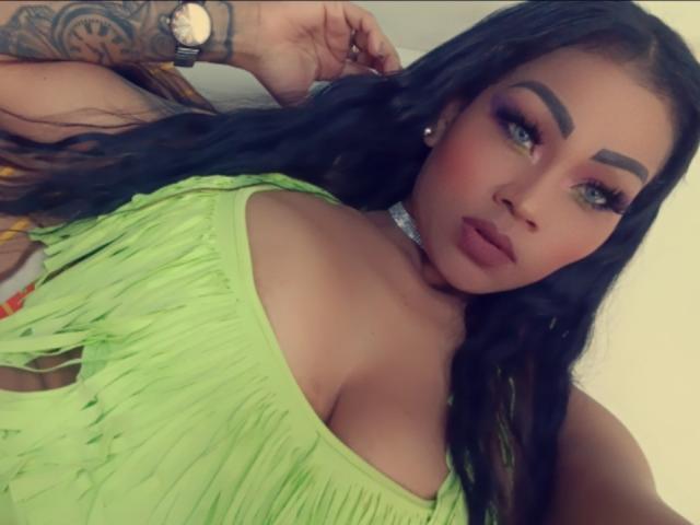 LinaaPrincess tem olhos verdes, sobrancelhas grossas e cabelos pretos longos. Ela está vestindo um vestido verde neon com franjas, que mostra seus seios grandes. Ela está olhando para a câmera..