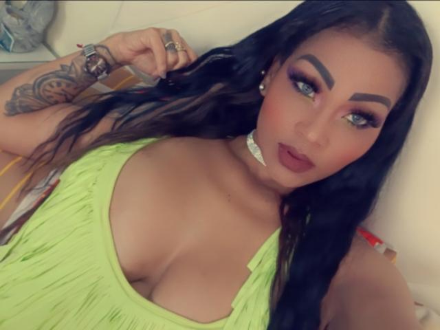 LinaaPrincess tem seios grandes, está vestida com um vestido verde neon, tem cabelo preto longo, olhos azuis, sobrancelhas marcadas, batom vermelho, um colar branco, uma tatuagem no braço e está olhando para a câmera..