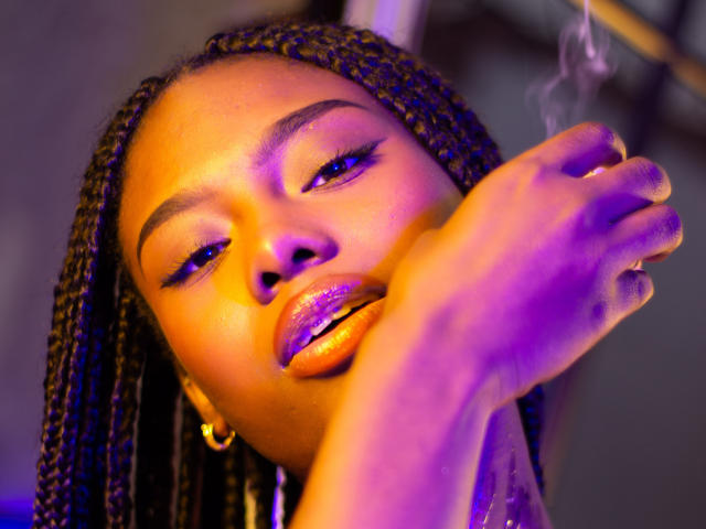 Ebony, met glanzende paarse lippen, houdt een smeulende wietjoint dicht bij haar gezicht, en ze ziet er ontspannen uit met gevlochten haar en gouden oorringen, verlicht door paarse en oranje lichten..