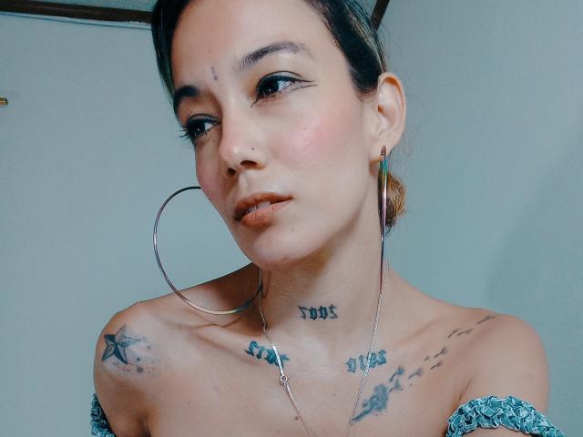 Veroo69 with tattoos on neck and shoulders, wearing off-shoulder blue top, hoop earrings, and nose ring, looking at camera with subtle smile, brown hair in bun.