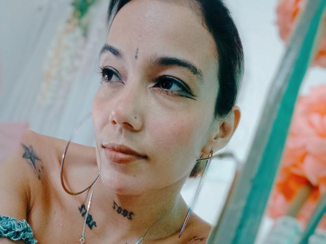 Veroo69 heeft donker haar, een neusring en oorringen. Ze draagt zwarte eyeliner en heeft tatoeages op haar borst en nek. Haar borsten zijn klein en ze glimlacht..
